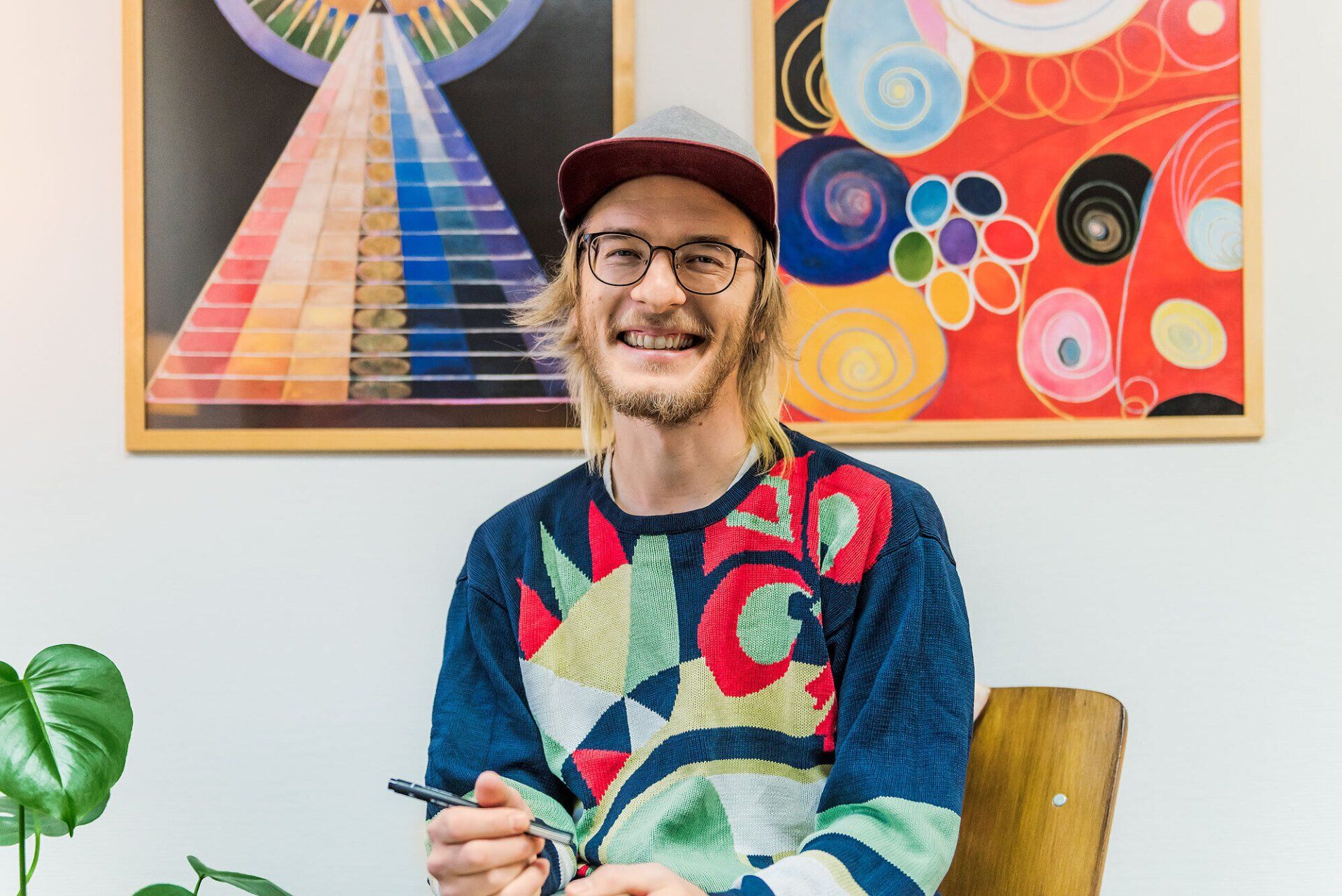 Poeten Jimmy Alm sitter framför två abstrakta, färgglada tavlor, iklädd keps och en mönstrad tröja. Han ler stort.